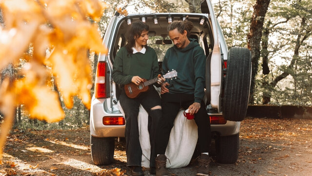 Couple sitting in car trunk in autumn forest, woman playing ukulele, man holding mug, enjoying cozy fall moment together