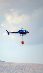 Firefighting helicopter flies with water bucket above wind turbine and forested hills