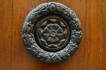 Close-up of an ornate, dark metal door knocker in the shape of a wreath with a central rosette design, against a textured wooden door