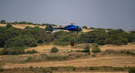 Firefighting helicopter flies with water bucket above wind turbine and forested hills