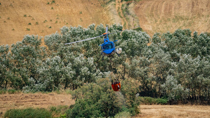 Firefighting helicopter flies with water bucket above wind turbine and forested hills