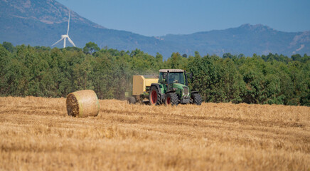 Obraz premium Modern tractor working in hay field during harvest, wind turbine and hills in background