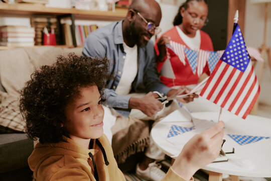 Child holding American flag while smiling, sitting at table with Black middle aged man and Black woman cutting paper flags, celebrating patriotic holiday indoors