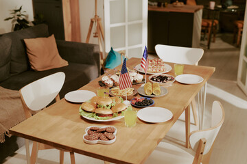 Table set with burgers, cupcakes, cookies, fruit, corn on cob, drinks and two American flags, suggesting festive meal preparation for holiday celebration in modern home dining area