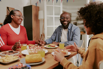 Black middle aged woman and Black middle aged man sitting at table smiling and talking with Black teenager while eating meal together, sharing food and enjoying family time indoors