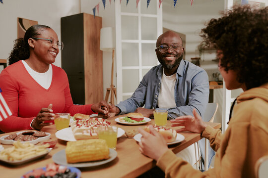 Black middle aged woman and Black middle aged man sitting at table smiling and talking with Black teenager while eating meal together, sharing food and enjoying family time indoors