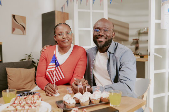 Black middle aged woman and Black middle aged man sitting together at table holding hands and smiling, woman holding American flag, celebrating with desserts and drinks indoors