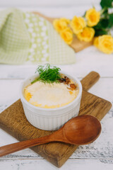 Baked casserole served in a white ramekin garnished with fresh green dill on a wooden board, accompanied by yellow roses and soft light.