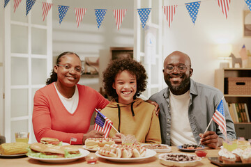 Black middle aged woman, Black middle aged man, and Black teenager sitting together at table holding American flags, smiling and celebrating with food and drinks during patriotic event