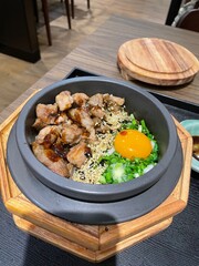 a traditional Korean stone pot filled with various ingredients. The hot pot contains rice, topped with savory grilled or stir-fried meat pork or vibrant green vegetable and a raw egg yolk