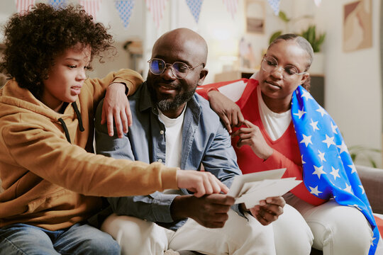 Black teenage boy sitting with middle aged Black man and middle aged Black woman draped in American flag, all looking at photographs together in living room decorated with flags