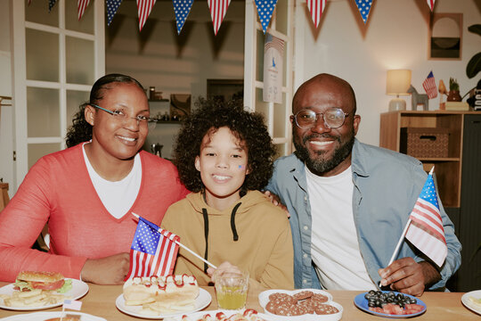 Portrait of Black woman, Black man, and child smiling and sitting together at table holding American flags, celebrating patriotic holiday with festive food and decorations