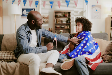 African American dad handing small American flag to child wrapped in United States flag, both...