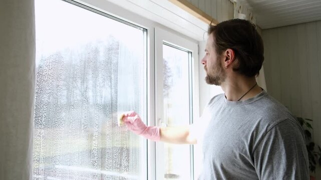 Man is cleaning window in apartment. Adult Male cleaner using a squeegee to wash a window. Bearded guy wiping and washing glasses with with spray bottle, rag and vacoom. Everyday chores. Purification.