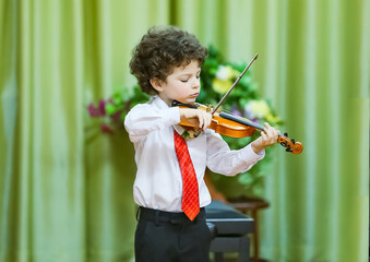 An eight-year-old boy plays the violin at a school concert. He is wearing a white shirt, dark trousers and a tie. He is standing on the stage, holding a violin and a bow, a background curtain