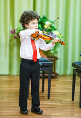 An eight-year-old boy plays the violin at a school concert. He is wearing a white shirt, dark trousers and a tie. He is standing on the stage, holding a violin and a bow, a background curtain