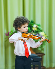 An eight-year-old boy plays the violin at a school concert. He is wearing a white shirt, dark...