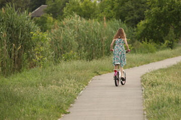 A girl rides a bicycle on an asphalt avenue among the greenery of the park. Active fun outdoors. Sports load for the proper development of the girl's body.