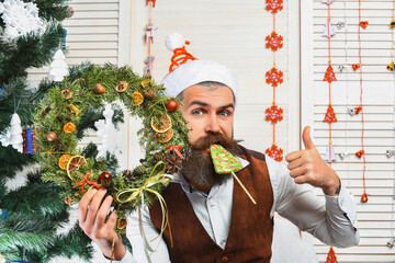 Xmas decoration for home. Christmas man eat candy. Bearded man at Christmas. New Year holiday. Man with moustache hold Xmas wreath. Happy New Year. Christmas wreath decoration and treats. Thumb up