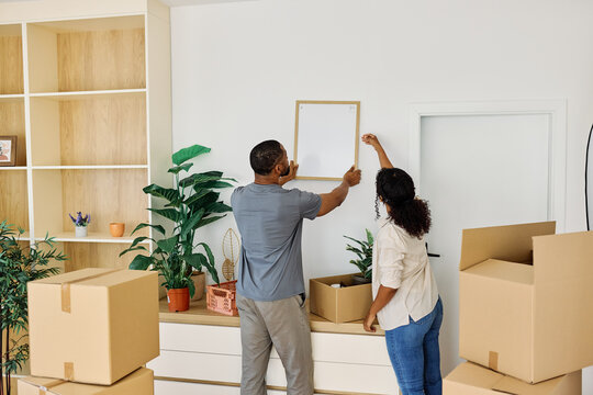 Portrait of a young couple unpacking, moving in and relocation to a new apartment, hanging a picture frame on wall, happy young couple family new beginning