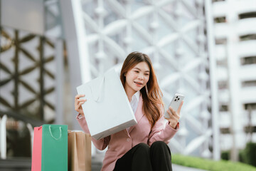 Happy young Asian woman shopping in a modern city during the evening, carrying bags, smiling,...