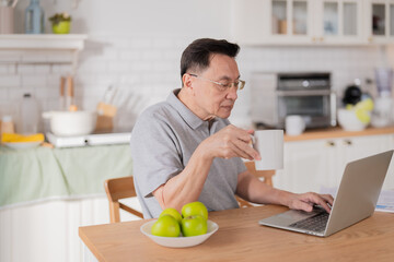 Mature senior man with glasses sits at the kitchen table in the morning, enjoying coffee while reading news updates on his laptop. With natural daylight streaming in, the gray-haired gentleman starts 