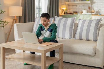 smart and cheerful Asian boy studies at home, typing on his computer and using a smartphone to...