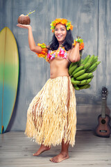 Ethnic cheerful woman in traditional Hawaiian costume holding a bunch of bananas and a coconut drink in an older studio with props