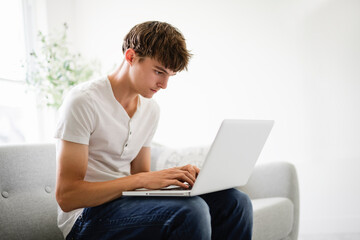 Handsome serious teenage boy sitting at home on the couch typing on laptop © Louis-Paul Photo