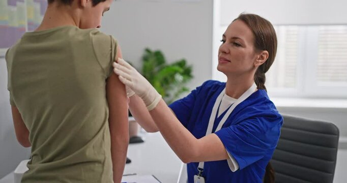 Female doctor in blue coat puts patch on vaccination site and gives candy to boy child after vaccination in hospital office