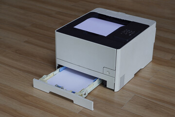 This photograph showcases a laser printer is on a wooden floor. The input and output trays loaded with white paper. The lower tray is extended, revealing a stack of paper ready for printing.