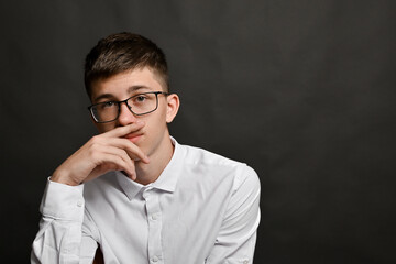 Thoughtful young man in glasses covers mouth with hand against gray studio background. Pensive...