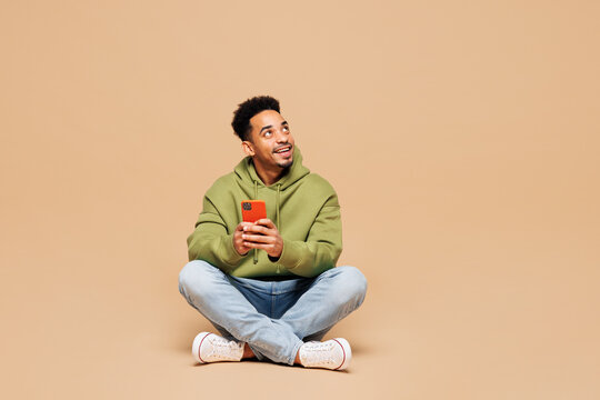 Fototapeta Full body young man of African American ethnicity wears green hoodie casual clothes sit hold in hand use mobile cell phone look aside isolated on plain pastel light beige background Lifestyle concept