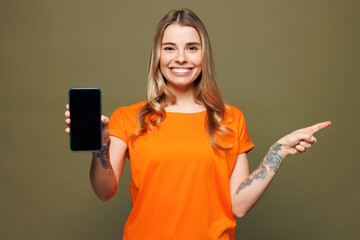 Young woman she wears orange t-shirt casual clothes hold in hand use mobile cell phone with blank screen workspace area point finger aside isolated on plain pastel green background. Lifestyle concept.
