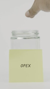 A yellow sticky note with the word opex is attached to a glass jar as a hand inserts a coin. This vertical video reflects saving for recurring business operations and costs
