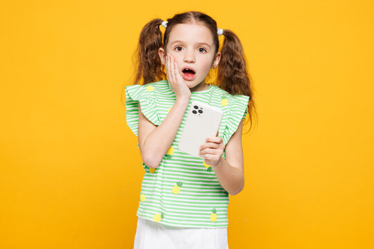 Little sad kid child girl 8-9 years old wears green t-shirt hold in hand use mobile cell phone cover mouth look camera isolated on plain yellow background. Mother's Day love family lifestyle concept.
