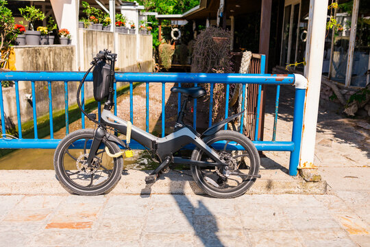 Compact electric bike parked by blue fence on sunny promenade. Urban mobility, eco transportation, and modern city lifestyle theme