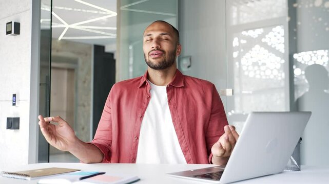 Businessman meditating with closed eyes during a break while sitting at workplace in business office. Calm worker practices yoga while breathing deeply, feels peace of mind, relaxes to relieve stress