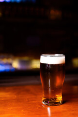 A glass of dark beer with a creamy foam stands on a wooden table in the center of the frame. The bar area is bathed in soft red light, creating a cozy and intimate atmosphere