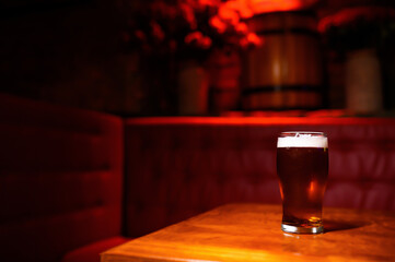 A glass of dark beer with a creamy foam stands on a wooden table in the center of the frame. The bar area is bathed in soft red light, creating a cozy and intimate atmosphere