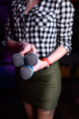 A woman in a checkered shirt and skirt holds three colorful microphones pointed toward the camera. Concept of karaoke, performance, or musical event. Focus on the microphones with a blurred background
