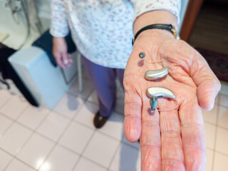 Elderly person holding a pair hearing aids in hand. assistive hearing technology in real-life setting, highlighting accessibility, senior healthcare, hearing loss. Bathroom, emphasizing daily use and 