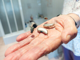 Elderly person holding a pair hearing aids in hand. assistive hearing technology in real-life setting, highlighting accessibility, senior healthcare, hearing loss. Bathroom, emphasizing daily use and 