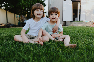Fototapeta premium Two smiling boys sitting on green grass, enjoying a sunny day together, showing the joy of childhood and friendship in a cozy outdoor setting.