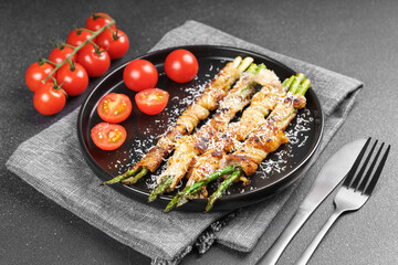 Grilled asparagus wrapped in crispy bacon sprinkled with parmesan cheese served on plate with fresh cherry tomatoes on black background. Gourmet dish, cooking recipe, cuisine, or restaurant menu