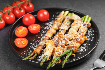 Grilled asparagus wrapped in crispy bacon sprinkled with parmesan cheese served on plate with fresh cherry tomatoes on black background. Gourmet dish, cooking recipe, cuisine, or restaurant menu