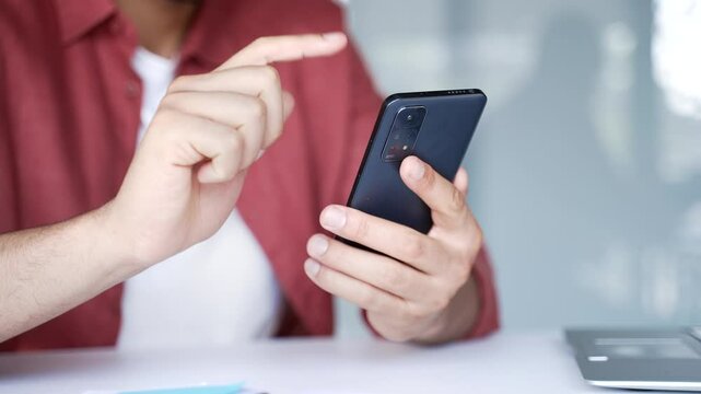 Close up of male hands holding mobile phone at desk at workplace in modern business office. Businessmen in red casual shirt using smartphone working, typing message chatting or swiping browsing online