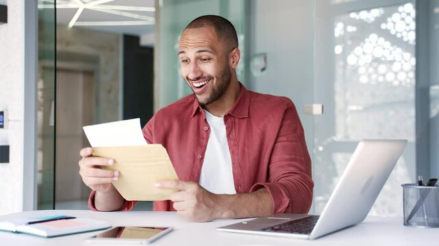 Smiling surprised businessman reading letter with great news sitting at workplace in modern business office. Joyful handsome worker is celebrating success, happy to receive positive good notification