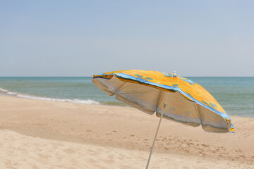 A yellow and blue beach umbrella stands on sandy shores, offering shade against the clear sky and...