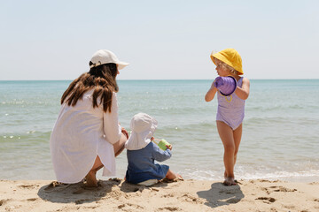 A mother and her two young children, facing the sea, enjoy a sunny day at the beach, with one child...